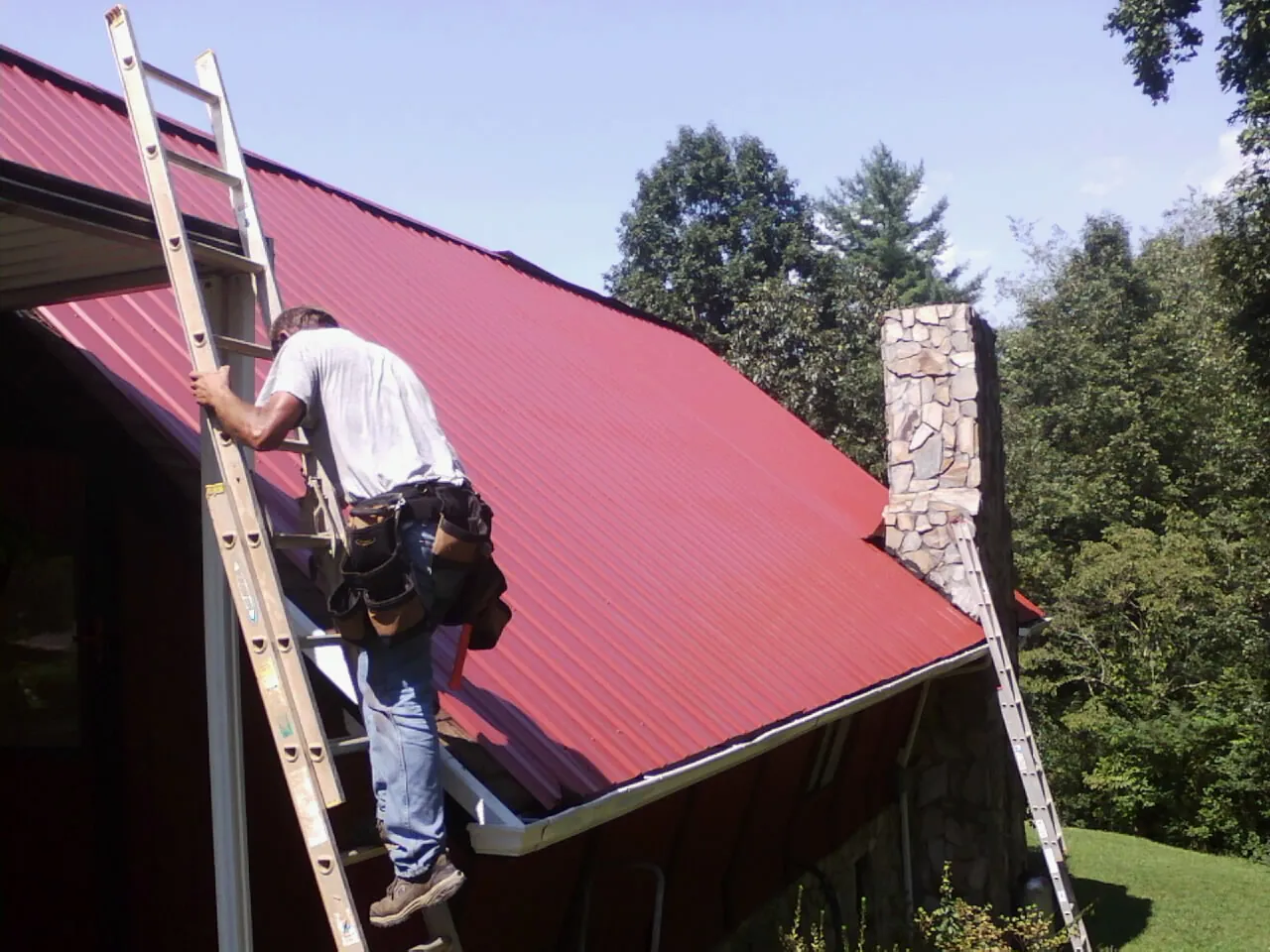 Roofer on a ladder beside a red metal roof and stone chimney — Soffit & Fascia Repair by Roofing Brothers in Big Cove Tannery